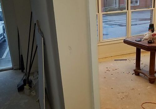 Renovation room with tools leaning by a doorway and debris on the floor near a table and windows.
