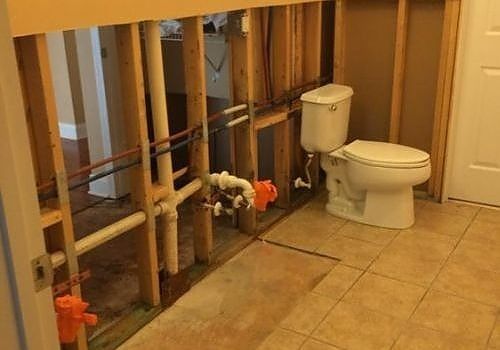 Exposed bathroom plumbing under a sink beside a white toilet and tan tile floor