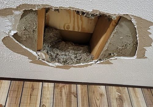 Damaged wall with a large hole exposing insulation and wooden studs above wood flooring