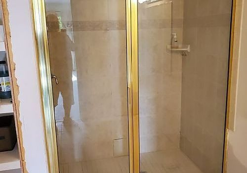 Glass shower enclosure with gold trim in a beige bathroom, with a person faintly reflected in the left panel