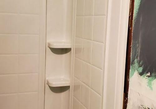 Corner shower with white tile and two built-in corner shelves, next to a dark painted wall.