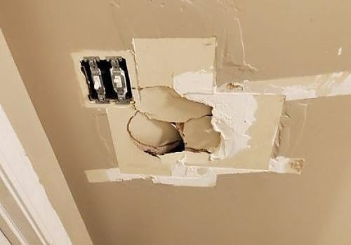 Damaged drywall with a large hole and peeled paint near an electrical outlet.