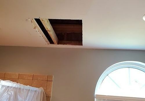 Bedroom ceiling with a large open hole exposing attic space near a window and bed