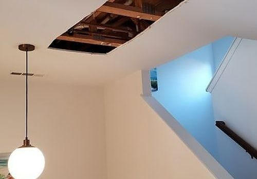 Ceiling with a large open hole exposing rafters, beside a stairwell with blue walls and a hanging light.