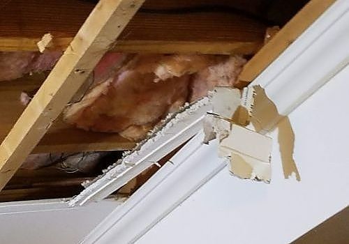 Broken white ceiling trim exposing wooden rafters and insulation above a wall.