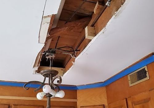 Damaged ceiling with a large hole exposing wood and wiring above a room with a hanging light fixture