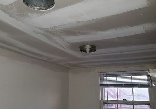 Unfinished ceiling with taped drywall seams and two light fixtures above a window with blinds