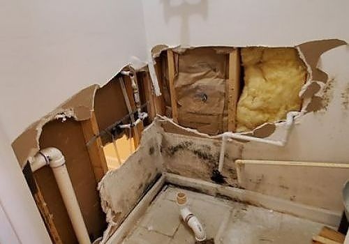 Bathroom wall torn open with exposed studs, insulation, and plumbing under a damaged sink area