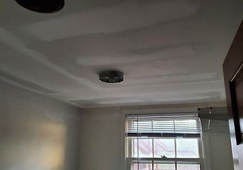 Ceiling with water damage and a stain near a window in a white room