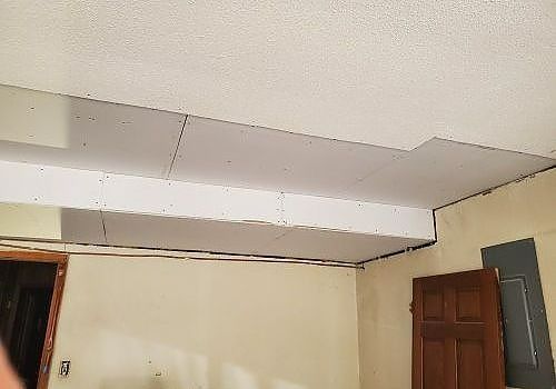 Indoor ceiling with a large damaged section and exposed beam above a beige wall and wooden door