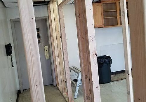 Renovation site with exposed wall studs, doorway, ladder, and black trash can in a hallway