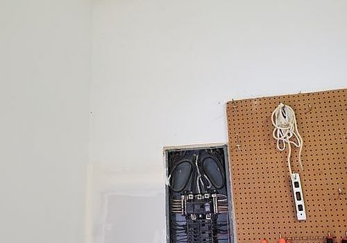 Wall with open electrical box and pegboard holding a coiled white cord and strip of outlets
