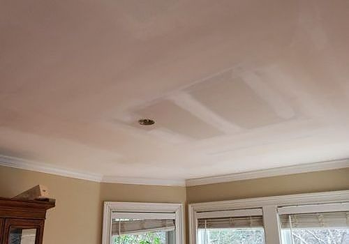 Living room ceiling with patchy drywall repairs above windows and beige walls