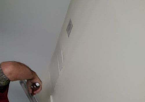 Hand holding a spray bottle near a plain white wall with a small vent or outlet.