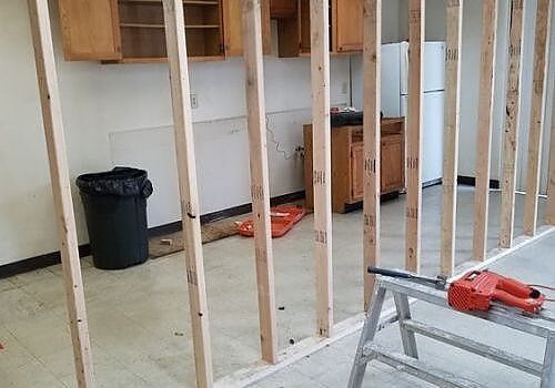 Unfinished room with exposed wall studs, a trash bin, and tools on a scaffold in the foreground.