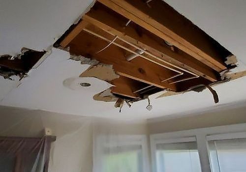 Ceiling with a large broken hole exposing wooden beams and debris above a room window.