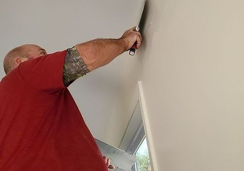 Person in a red shirt using a drill to install something on a white wall near a mirror.