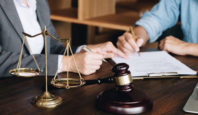 Scales of justice, gavel, and a person signing a document with another person present in an office.