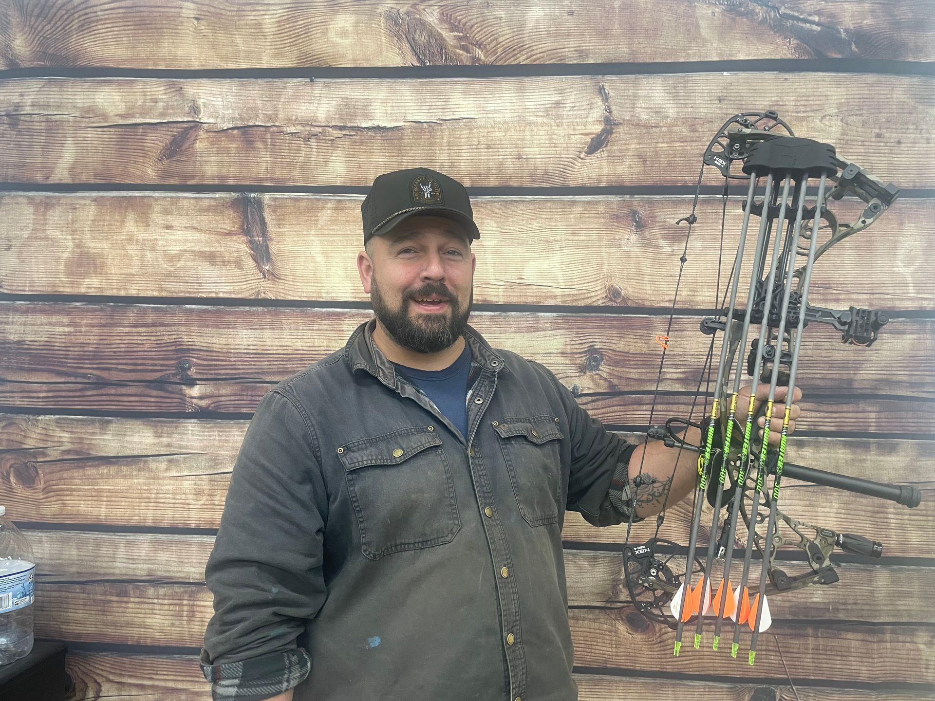Man holding a compound bow with arrows, standing in front of a wooden wall.