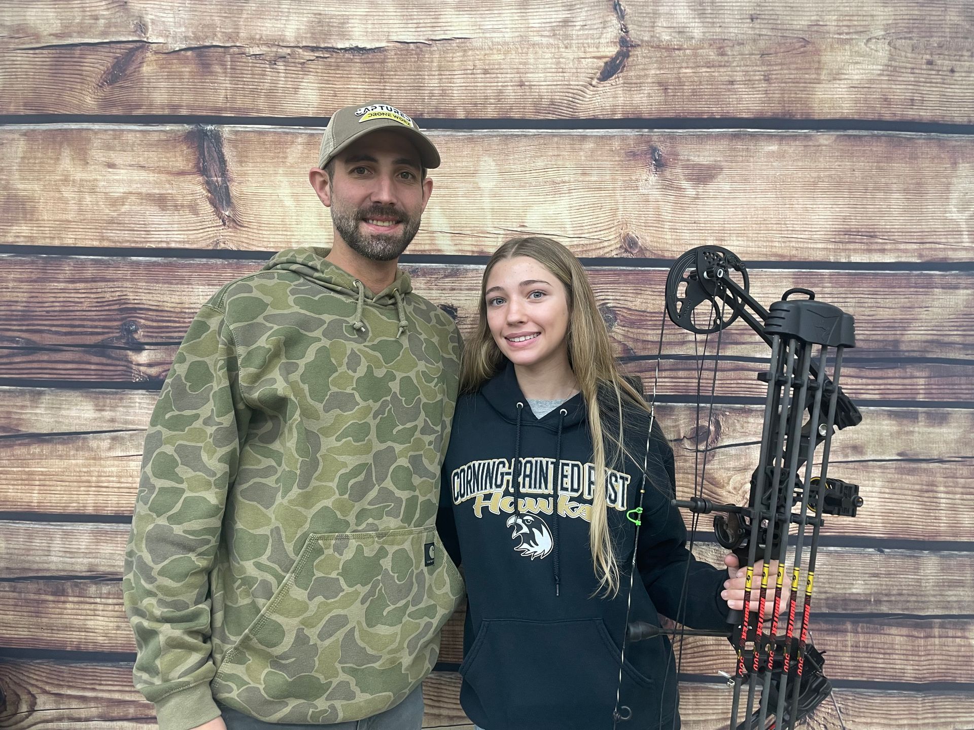 Two people standing in front of a wooden wall beside a compound bow, wearing camo and a hoodie.