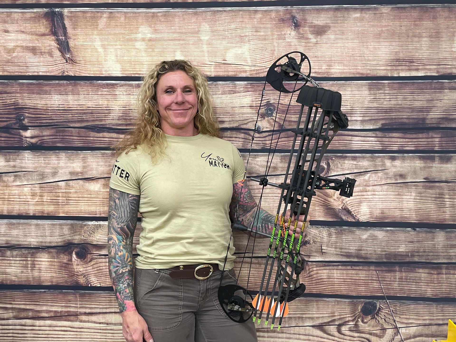Woman holding a compound bow, standing before a wooden plank backdrop, wearing a tan shirt and tattoo sleeves