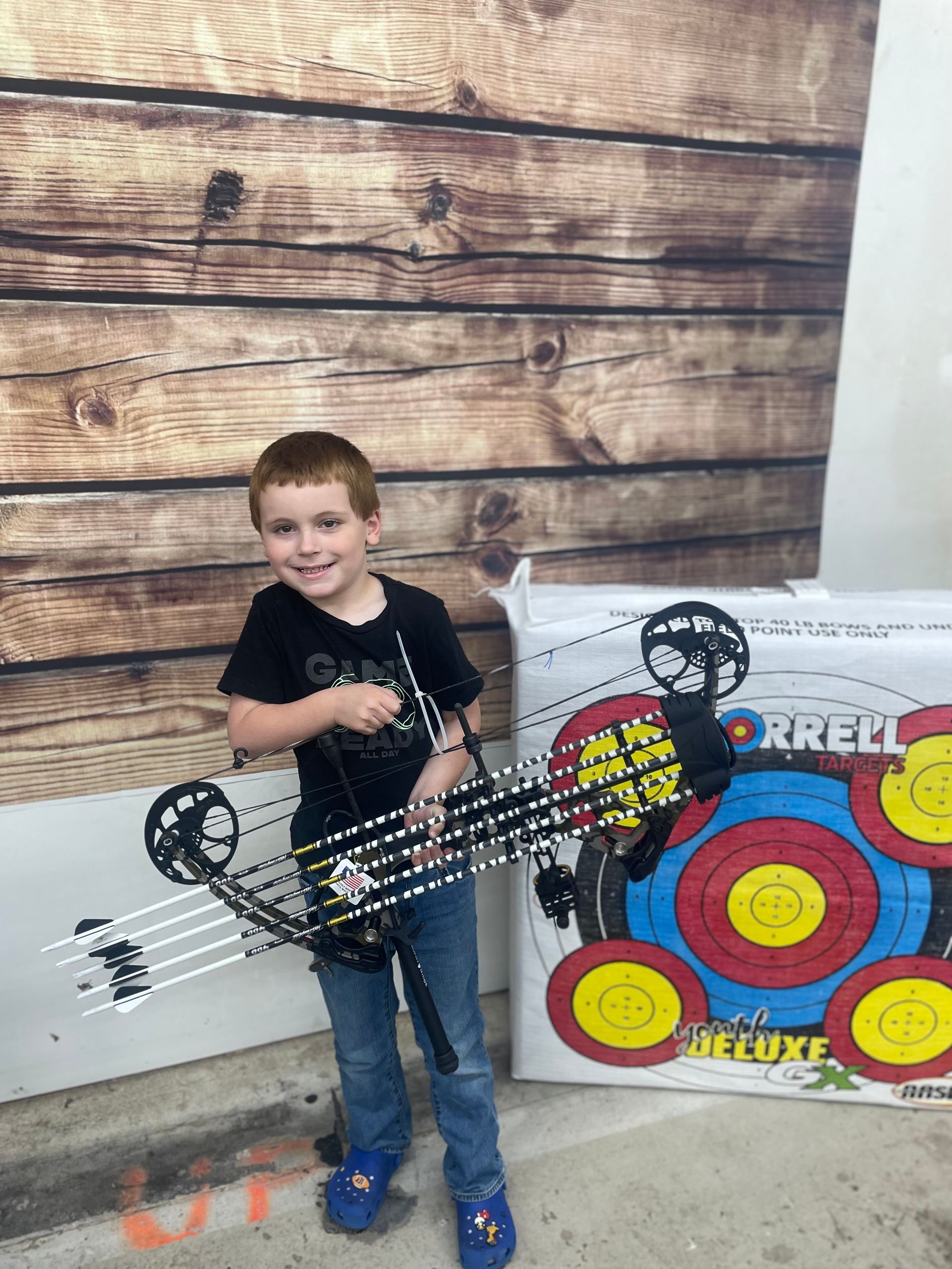 Boy standing by a wooden wall holding a toy crossbow beside a colorful target board