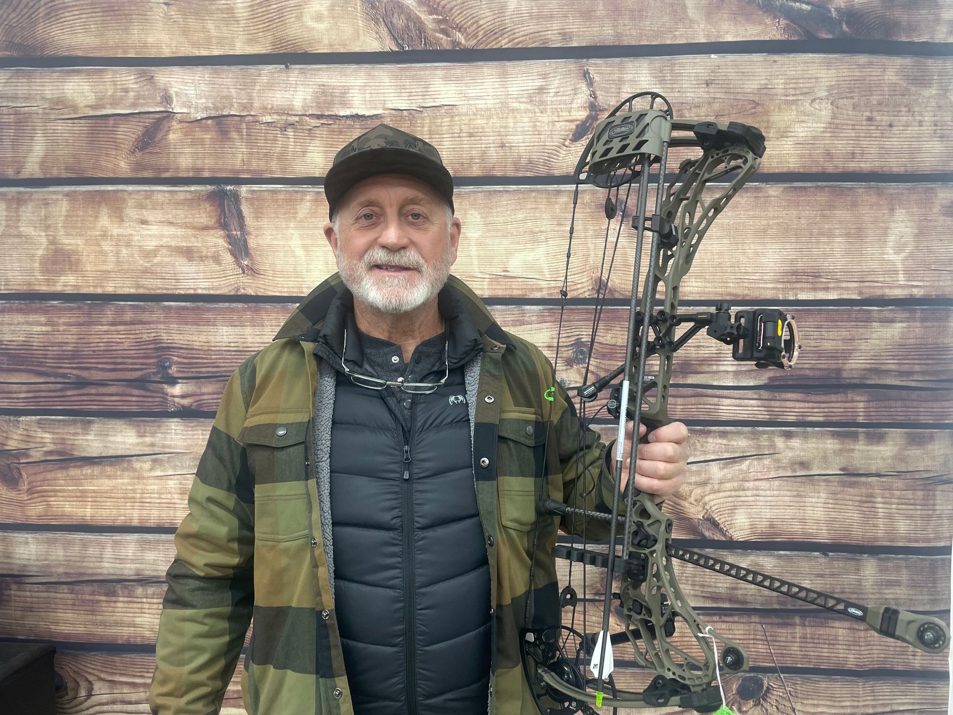 Man holding a compound bow, standing before a wooden wall, wearing a cap and camouflage jacket.