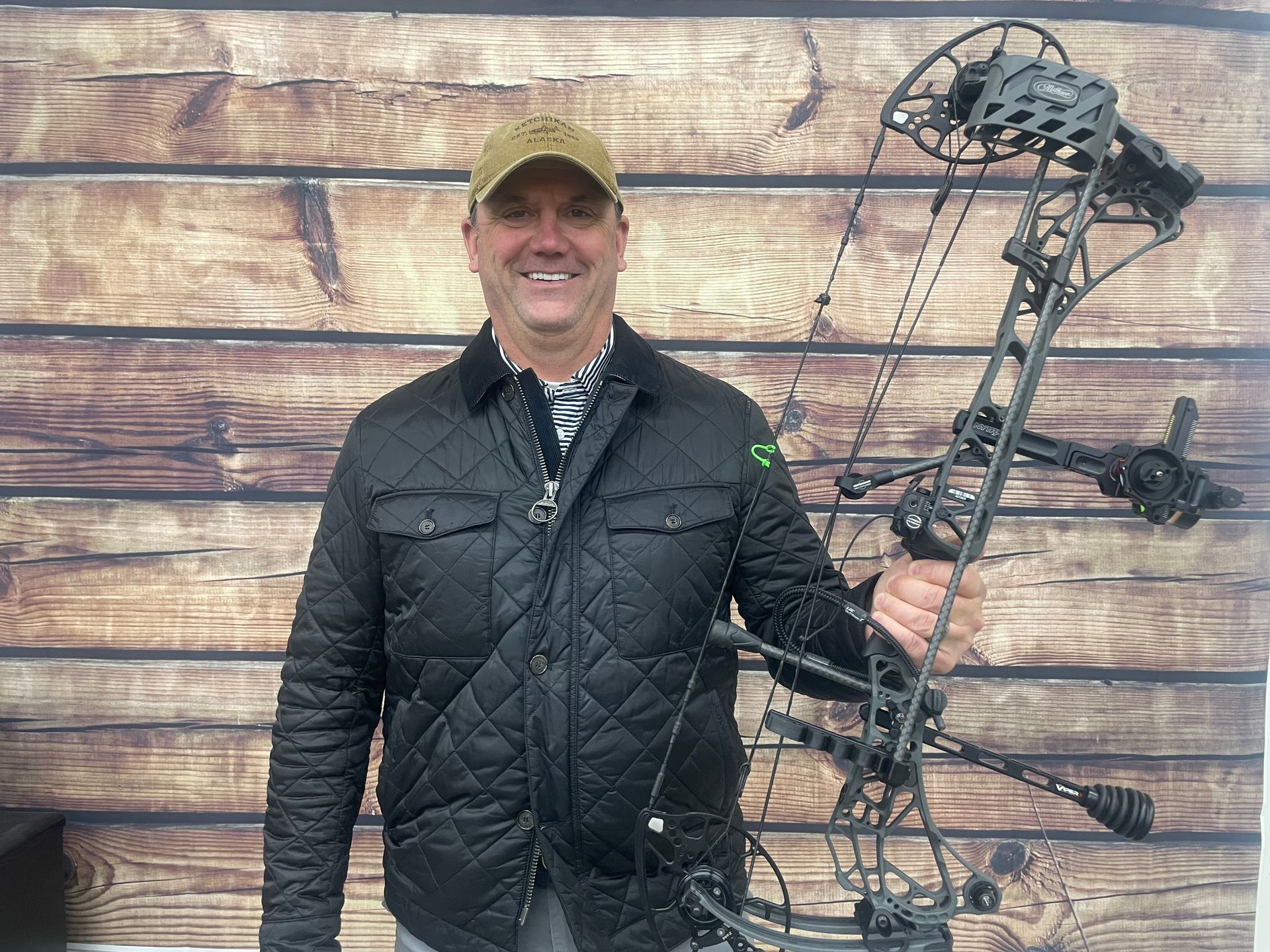 Smiling man holding a compound bow, standing in front of a wooden plank wall.