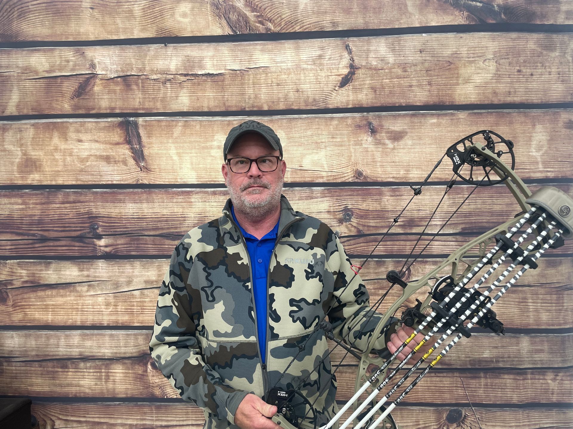 Man holding a compound bow in front of a weathered wooden wall, wearing camo jacket and cap.