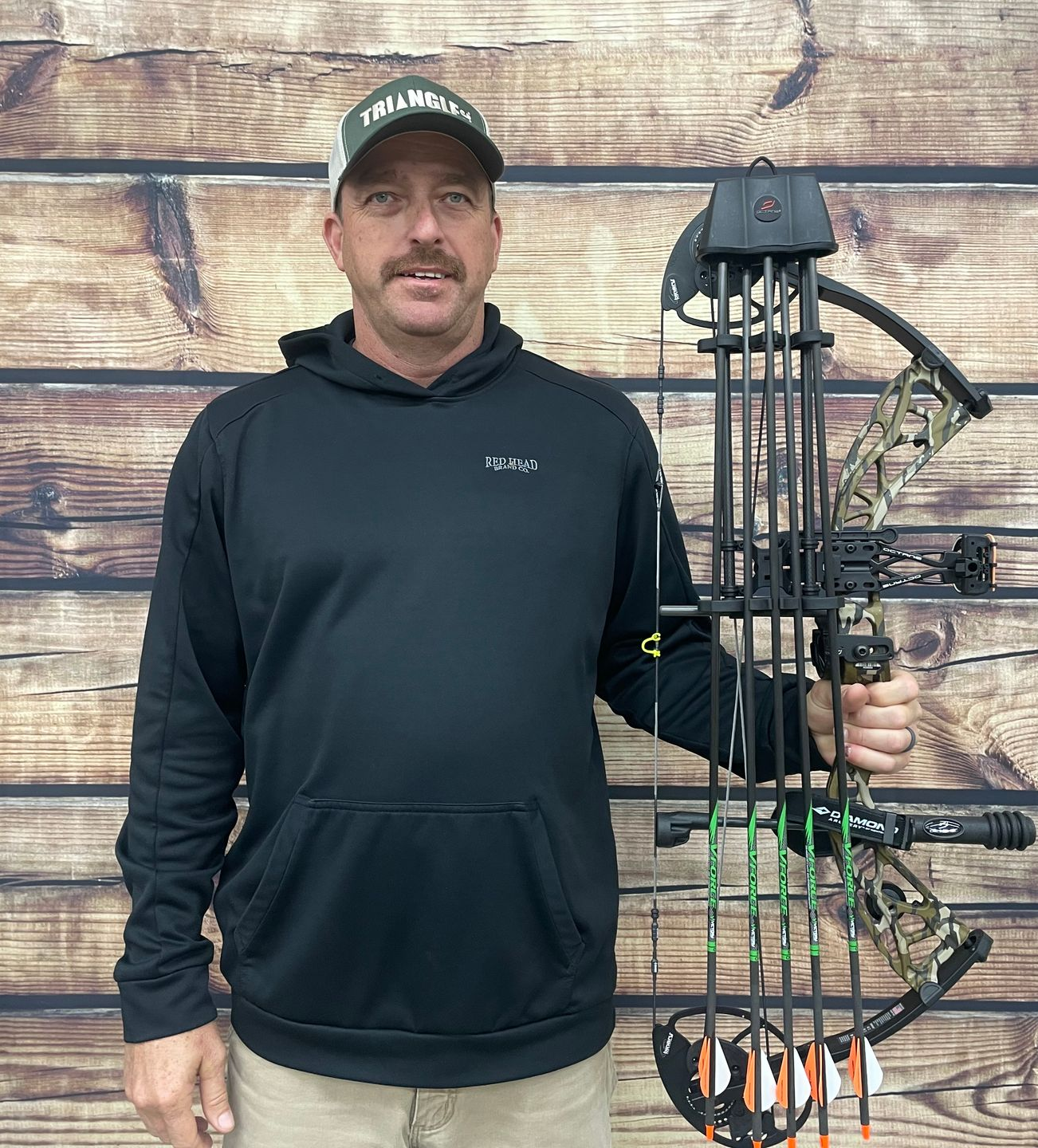 Man holding a compound bow, wearing a black hoodie and hat, standing in front of a wood background.