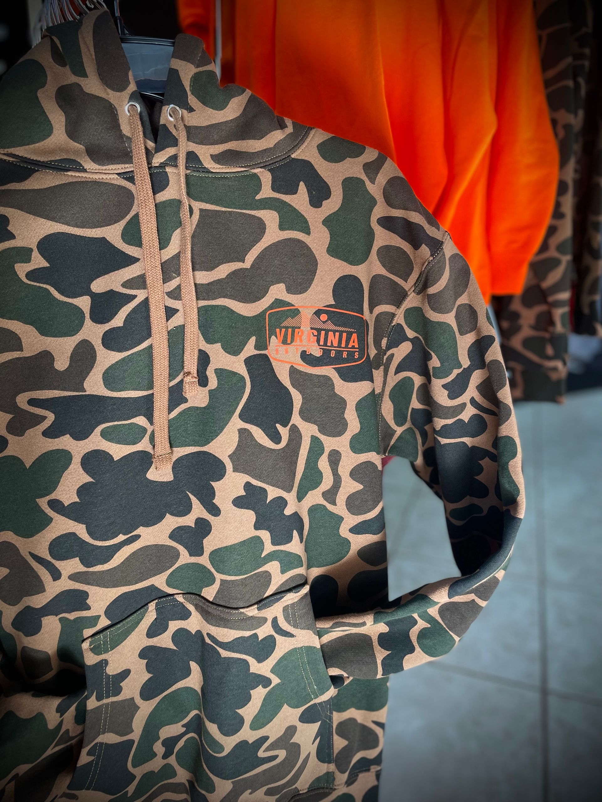 Camouflage hoodie with brown drawstring and logo, hanging in a store. Orange jacket visible in background.