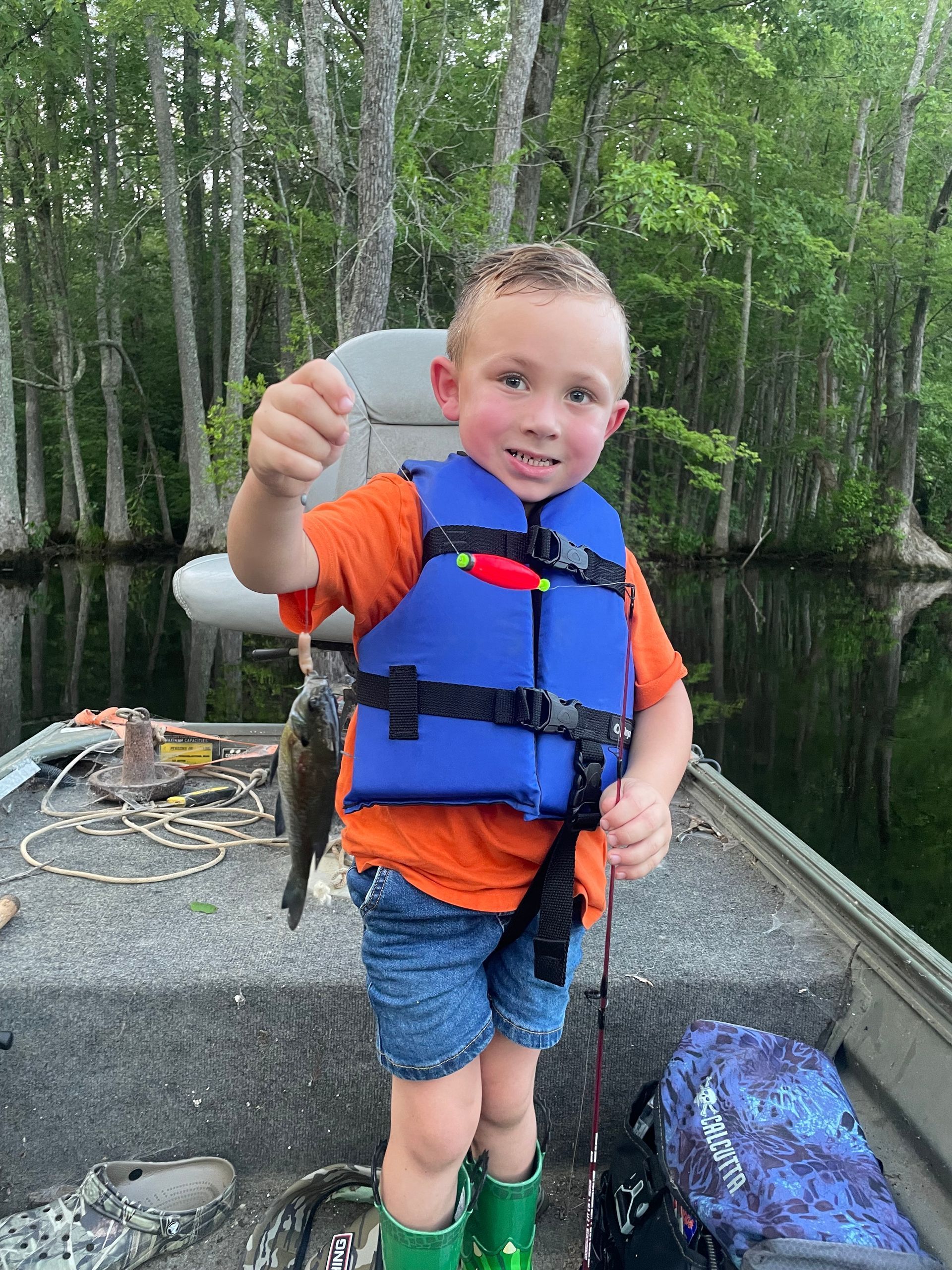 A young boy in a life jacket is standing on a boat holding a fish.