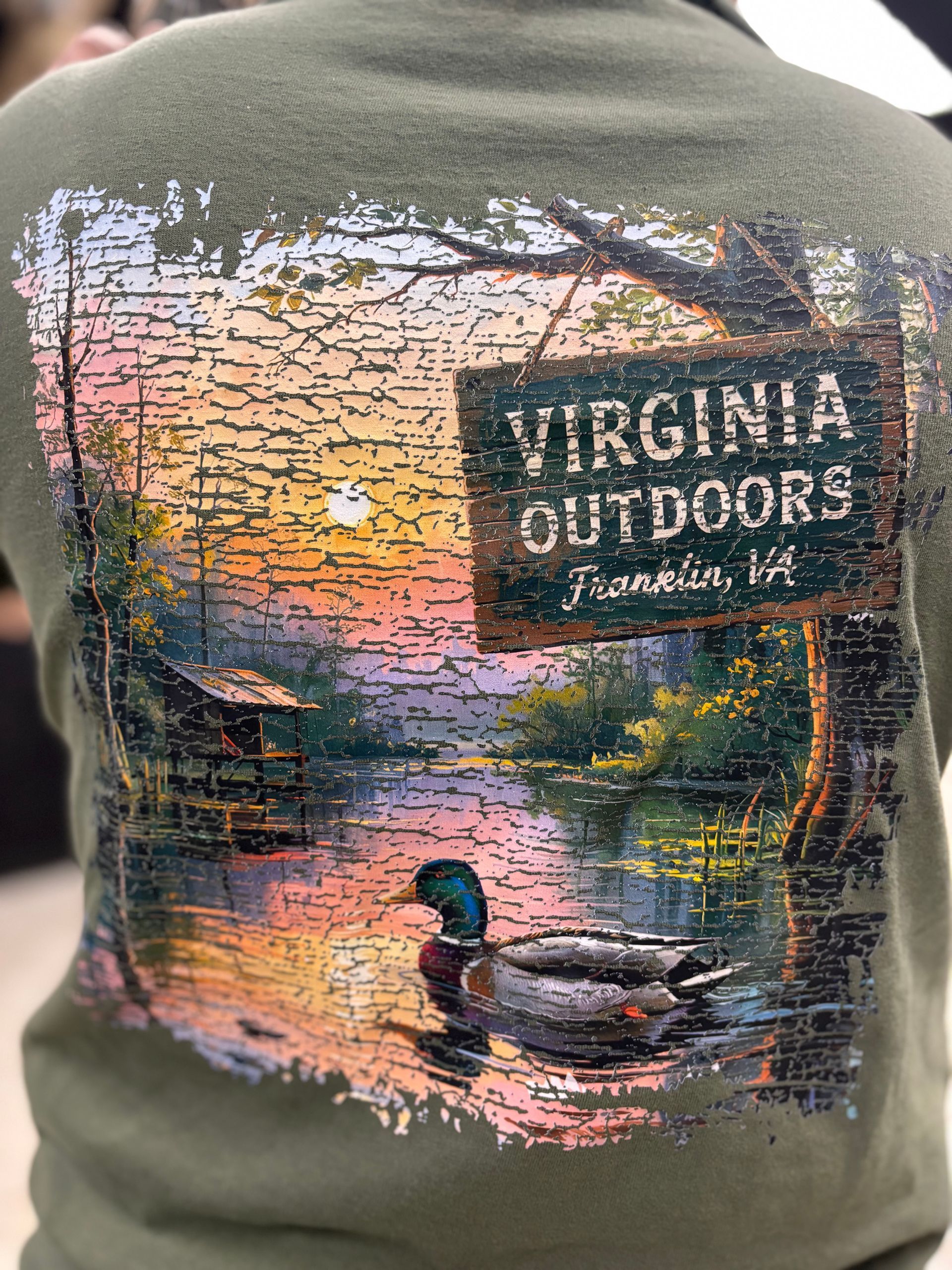 Back of a green t-shirt with a Virginia Outdoors sign and a duck in water at sunset.