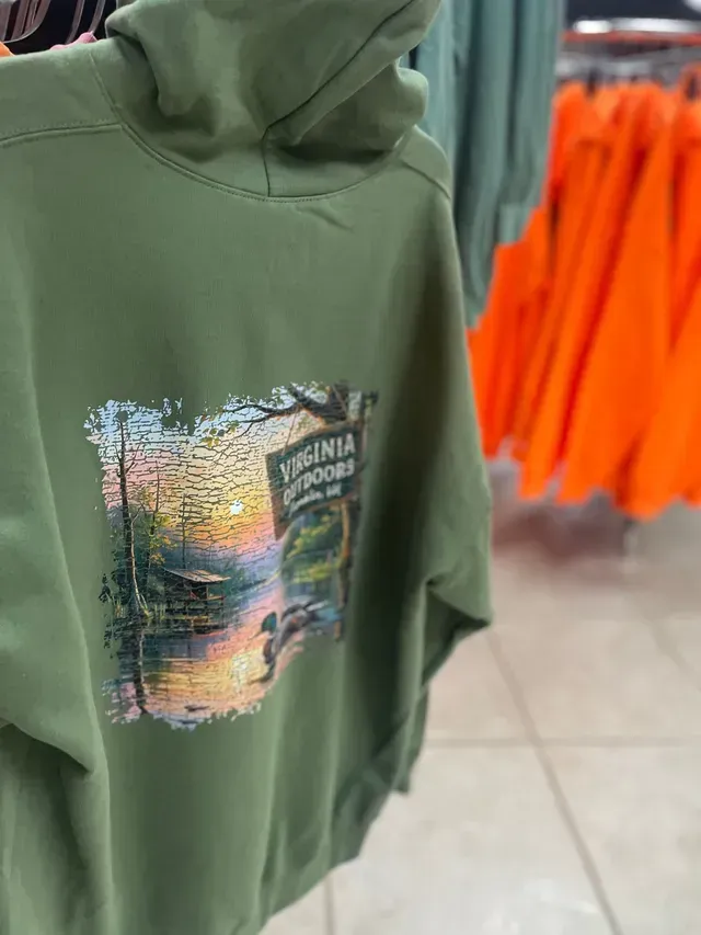 Green hoodie with a colorful forest graphic in a clothing store, orange shirts blurred in background