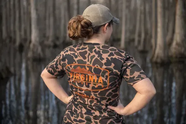 Person in camo shirt and cap, viewed from behind, standing with hands on hips.