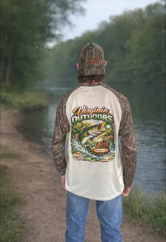 Person standing on a path by a river, wearing a camo cap and long-sleeve fishing shirt with a large fish graphic on the back