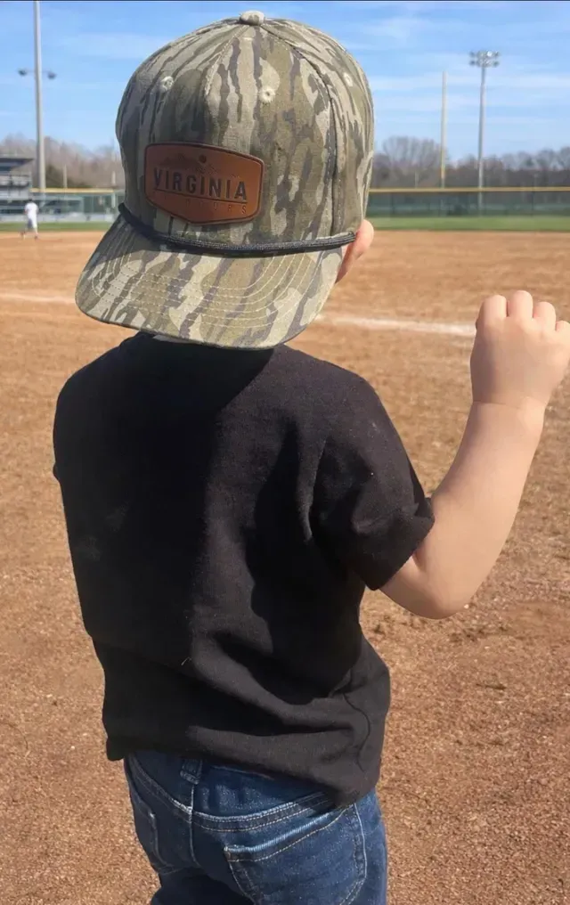 Child in a camouflage cap and black shirt standing on a baseball field, seen from behind.
