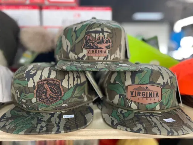 Camouflage Virginia baseball caps stacked on a shelf, with embroidered team and state patches