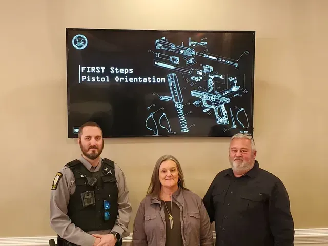 Three people stand in front of a screen showing “FIRST Steps Pistol Orientation” and a firearm diagram.