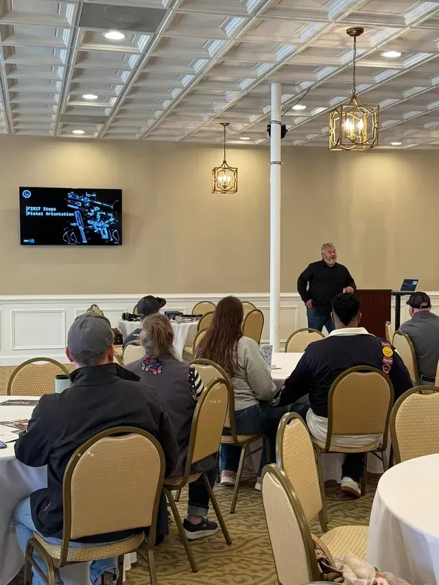 Conference room presentation with seated audience facing a speaker beside a wall-mounted screen.
