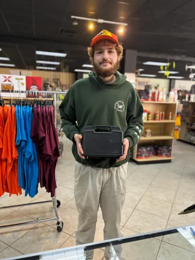 Man holding a black case in a store, wearing a green hoodie and orange hat.
