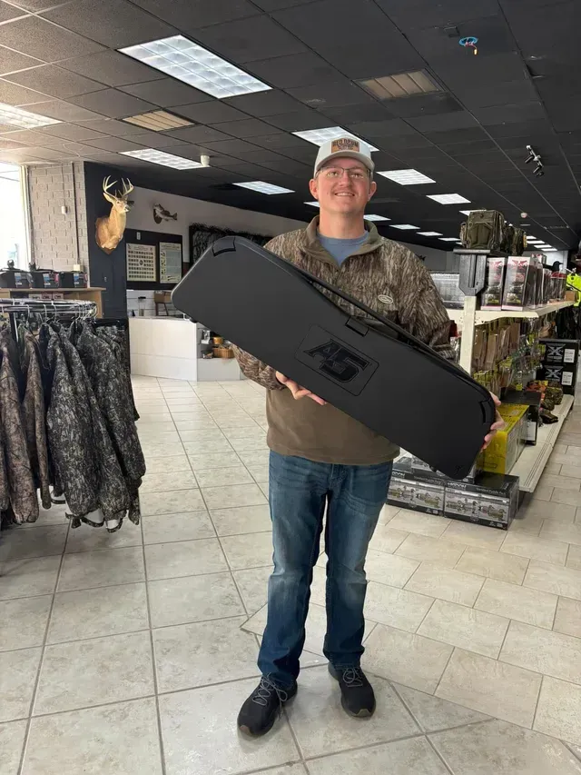 Man holding a black rifle case inside a store with hunting supplies, wearing a hat and camo jacket.