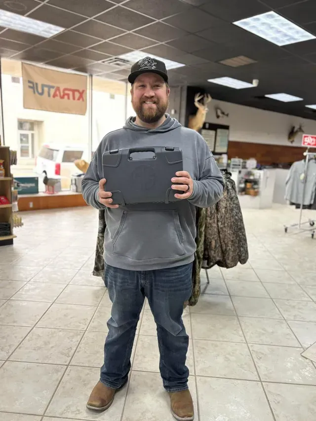Man in cap and hoodie holding a gray case in a store with hunting supplies.