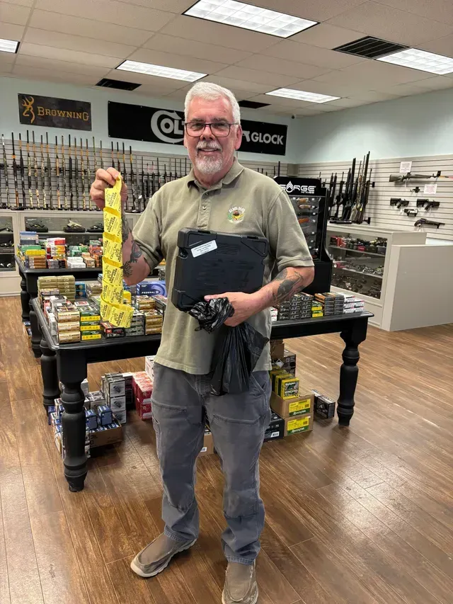 Man in a gun store holds a tie and black items. He is smiling. The setting has guns and displays.