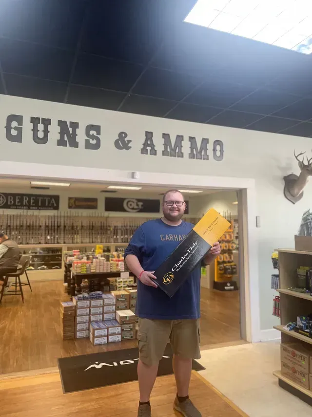 Man holding a black-and-yellow box in a guns and ammo store, with shelves and merchandise behind him.