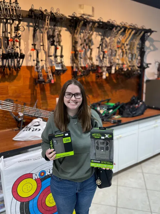 Person holding two black-and-neon-green packages in a gear store, with bows and arrows displayed behind.