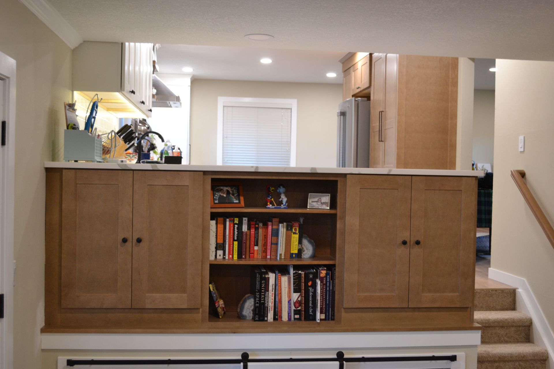 Built-in wooden bookcase with cabinets, open shelves, and a marble countertop, separating a kitchen from a lower level with stairs.