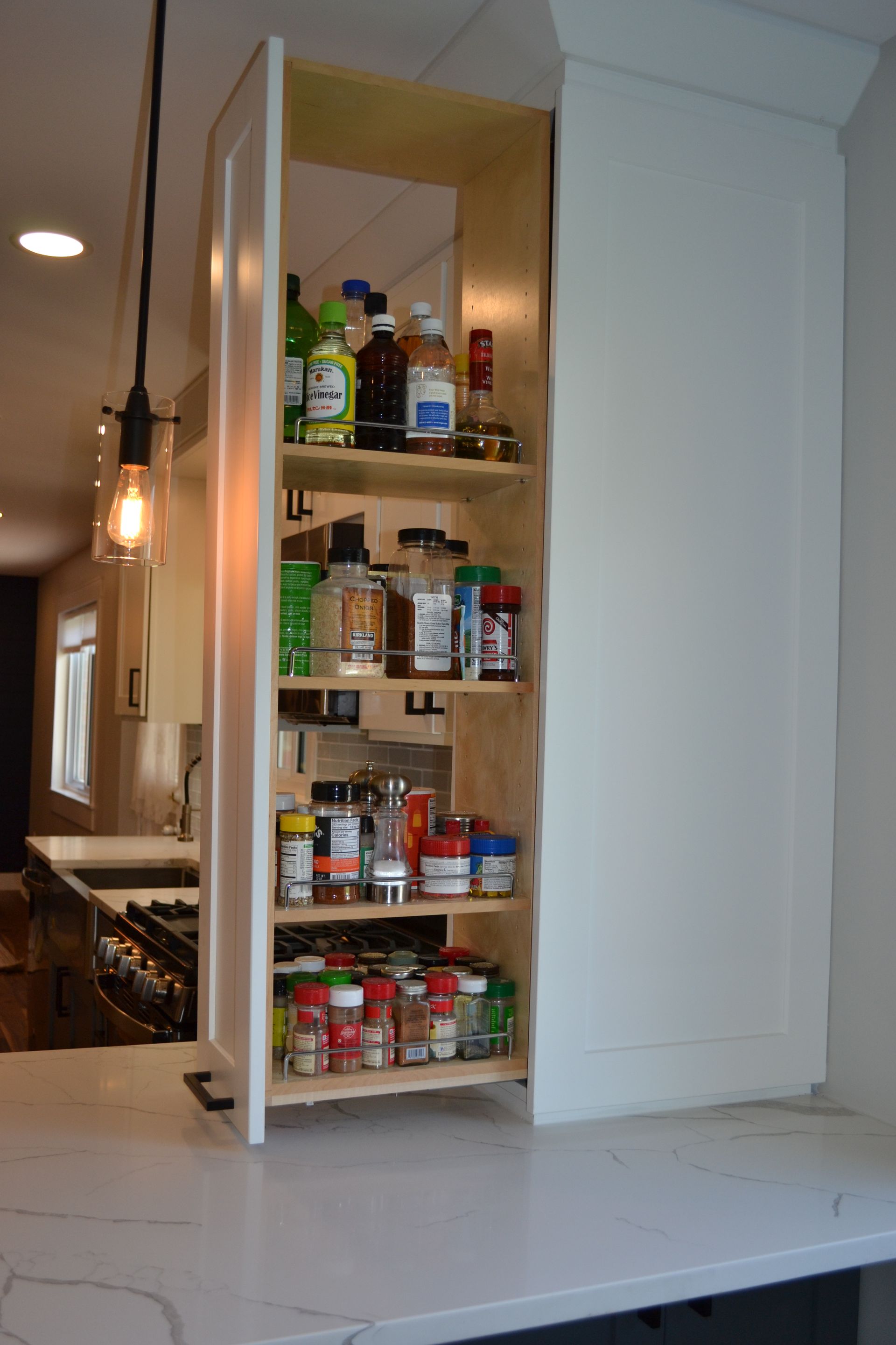 A tall, narrow pull-out kitchen cabinet filled with spices and bottles, set on a white countertop.