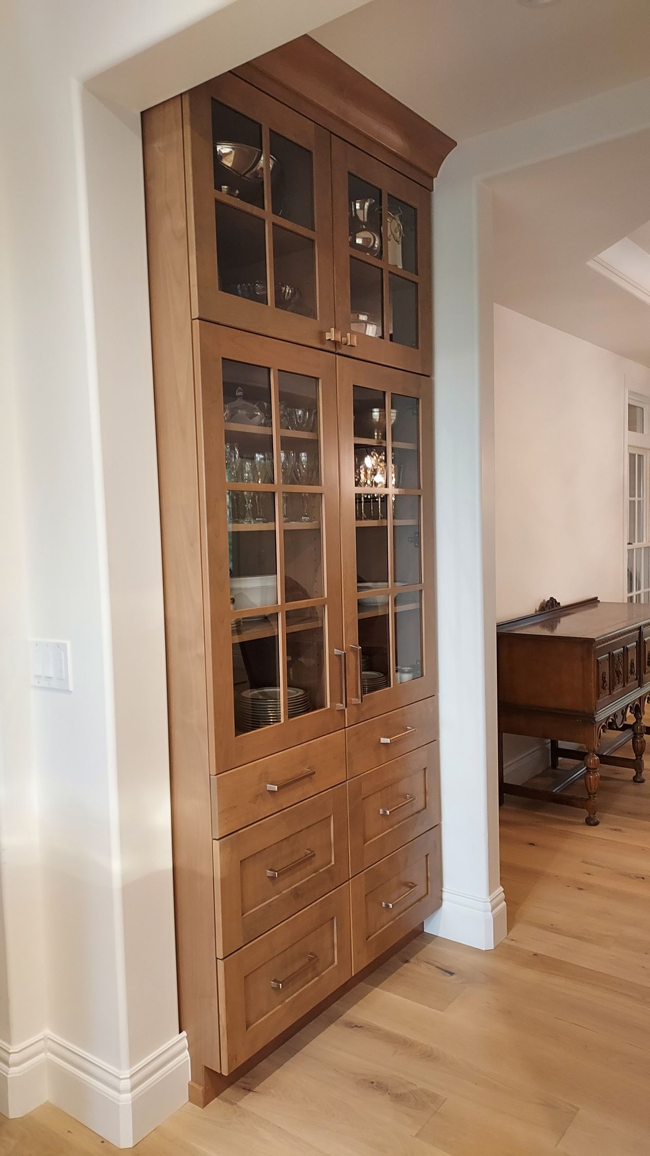 Tall, wooden built-in cabinet with glass doors and drawers, placed in a hallway.