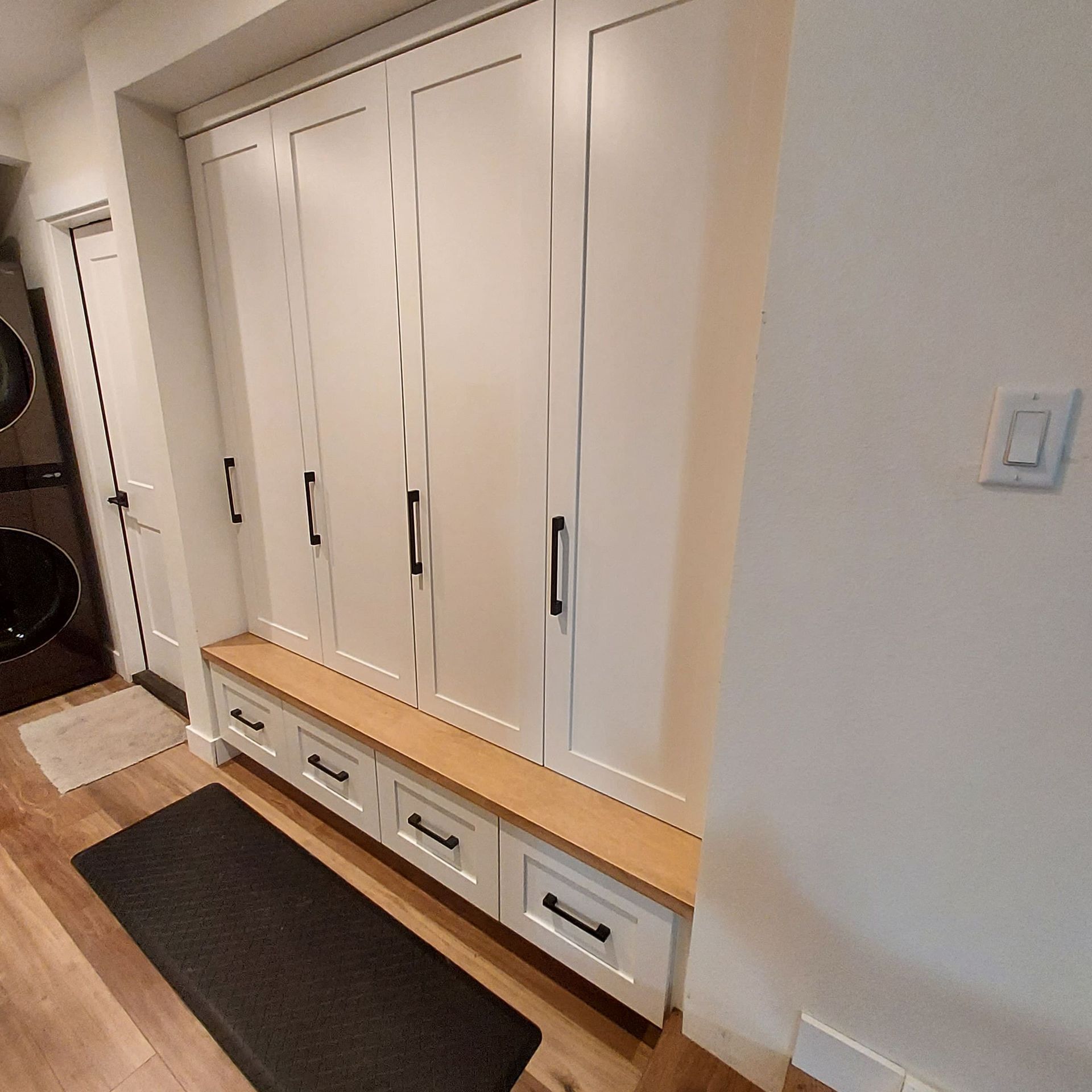 White built-in storage unit with cabinets and drawers, a wood bench, and a black mat in a hallway.